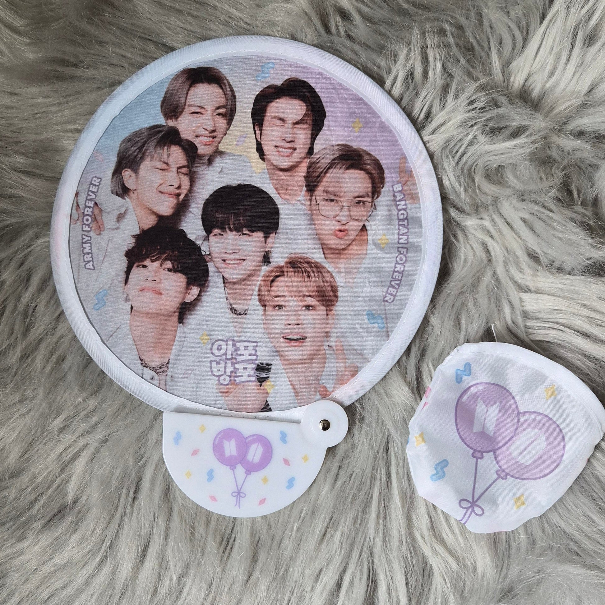 A round printed mirror or compact-style accessory with a group photo on the front, shown with two small matching accessories on a gray faux-fur surface. The design includes pastel purple balloon graphics and Korean text.