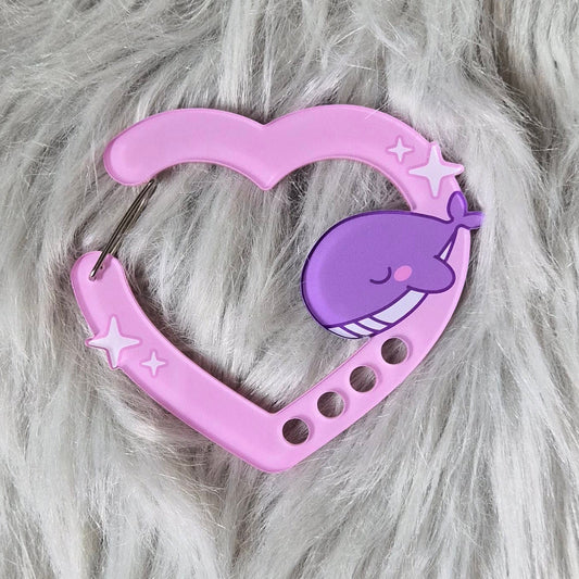 A pink heart-shaped silicone pet lick mat with a purple cartoon whale design, placed on a white faux fur background.